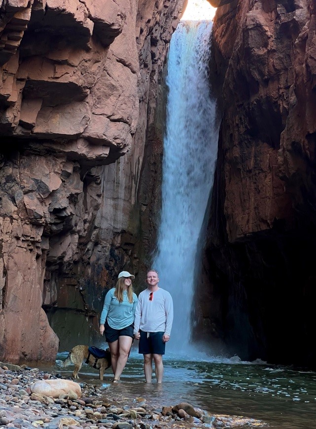 two people standing in the water with a dog in front of a waterfall
