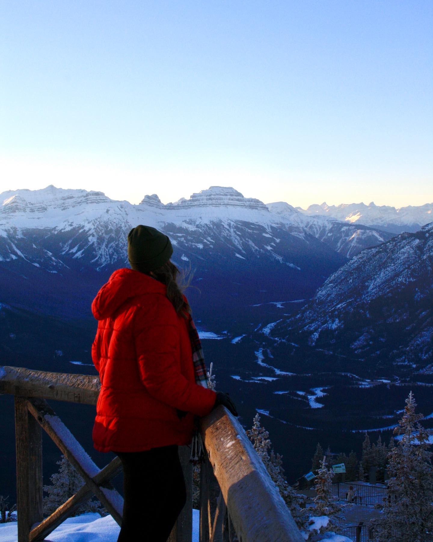 Girl in Red Coat Leaning on a Railing Overlooking Snow- Covered Mountains