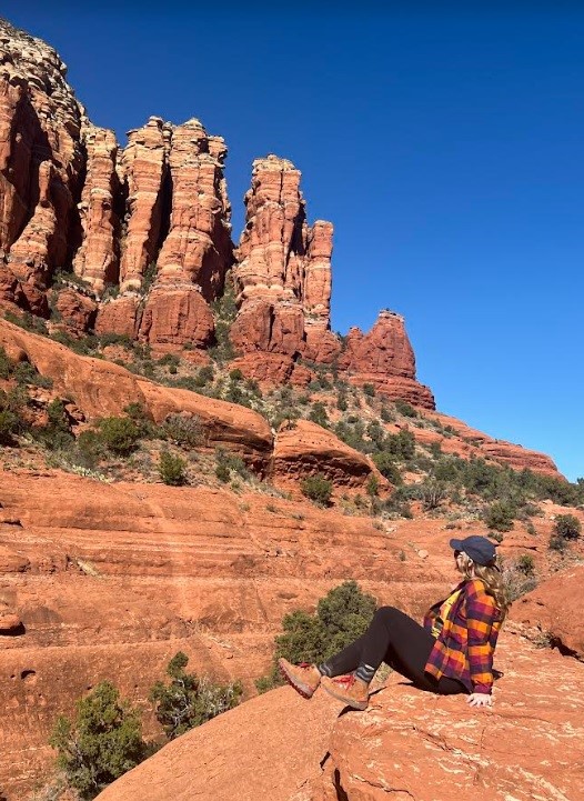 girl in pink plaid and dark pants sitting on red rock with a blue sky
