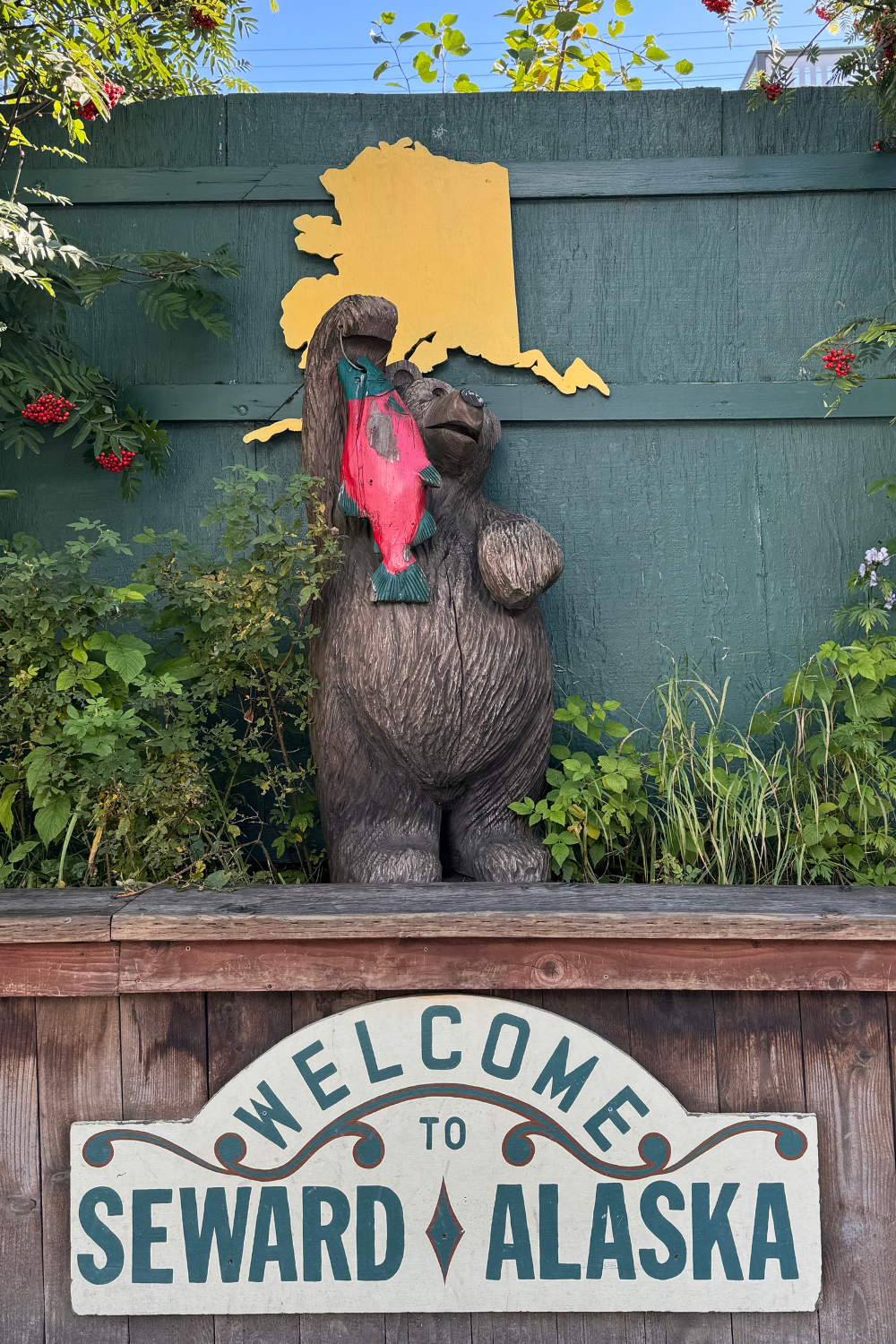 a bear holding salmon with a sign saying welcome to seward alaska