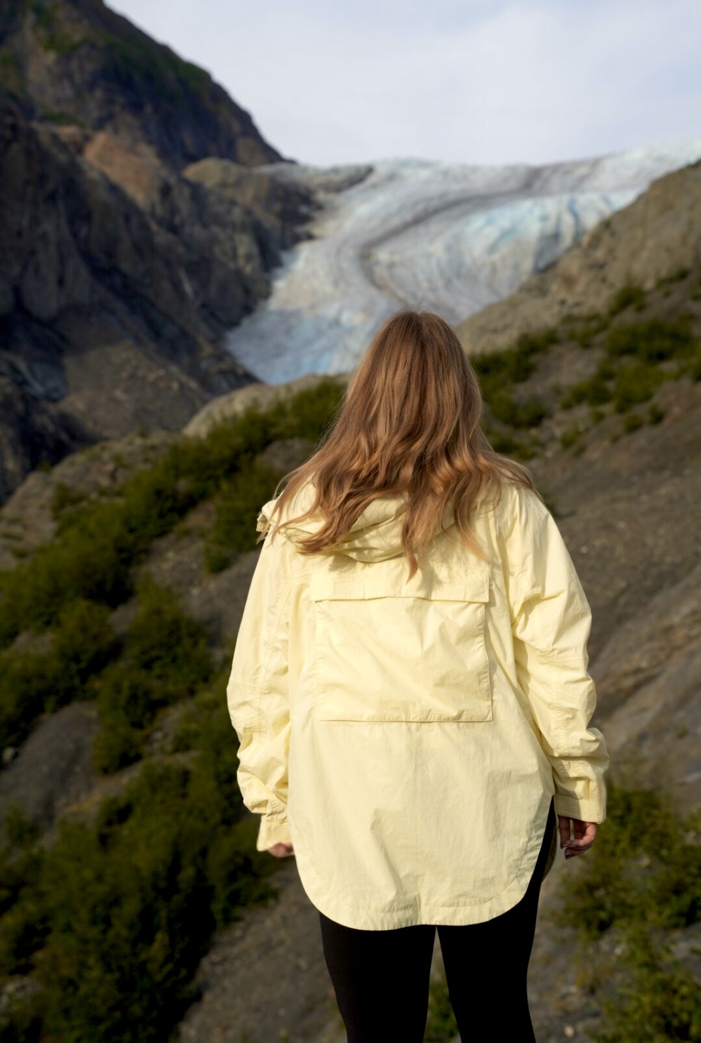 A person wearing a yellow jacket is standing on a rocky landscape, facing and looking towards a glacier and mountainous scenery.