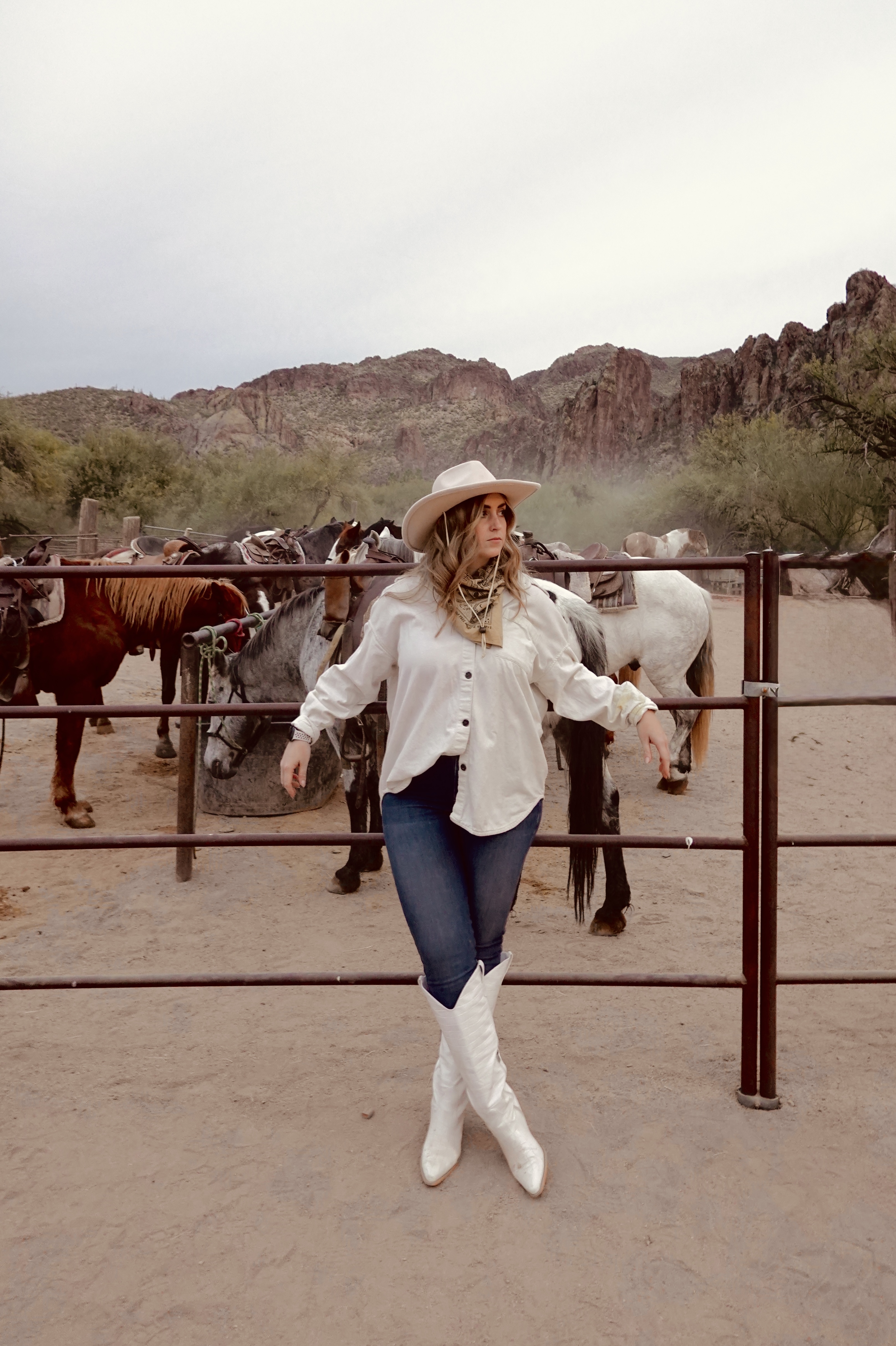 girl in jeans and tall white cowboy boots with a white shirt and white cowboy hat standing in front of a gate with horses