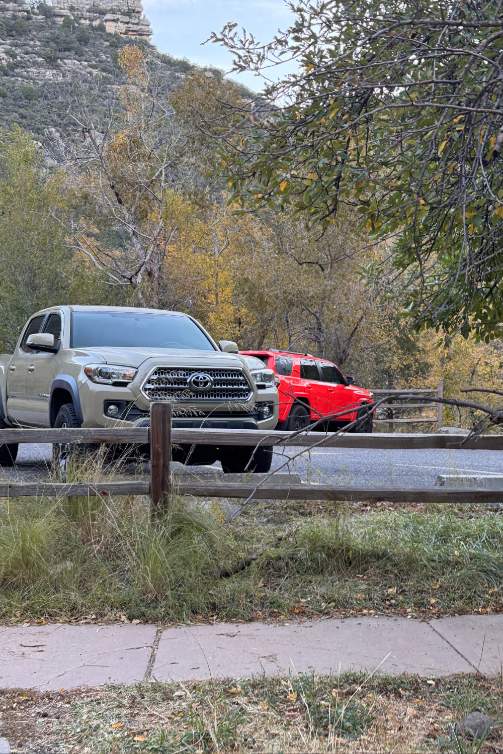 Two trucks, one gray and the other red, are parked by a roadside. Autumn trees and rocky cliffs form the scenic backdrop, conveying a tranquil outdoor vibe.