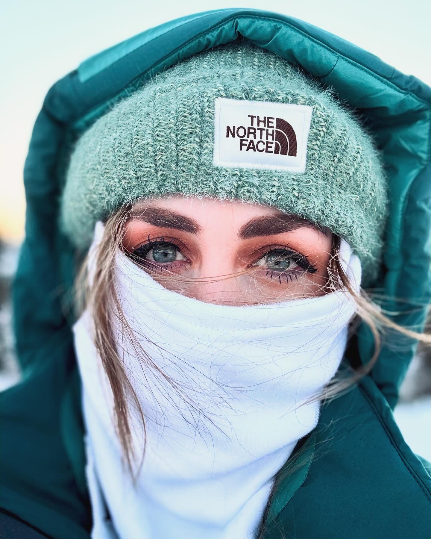 Close-up of a person with blue eyes, wearing a green beanie and teal hooded jacket, with face covered by a white cloth.