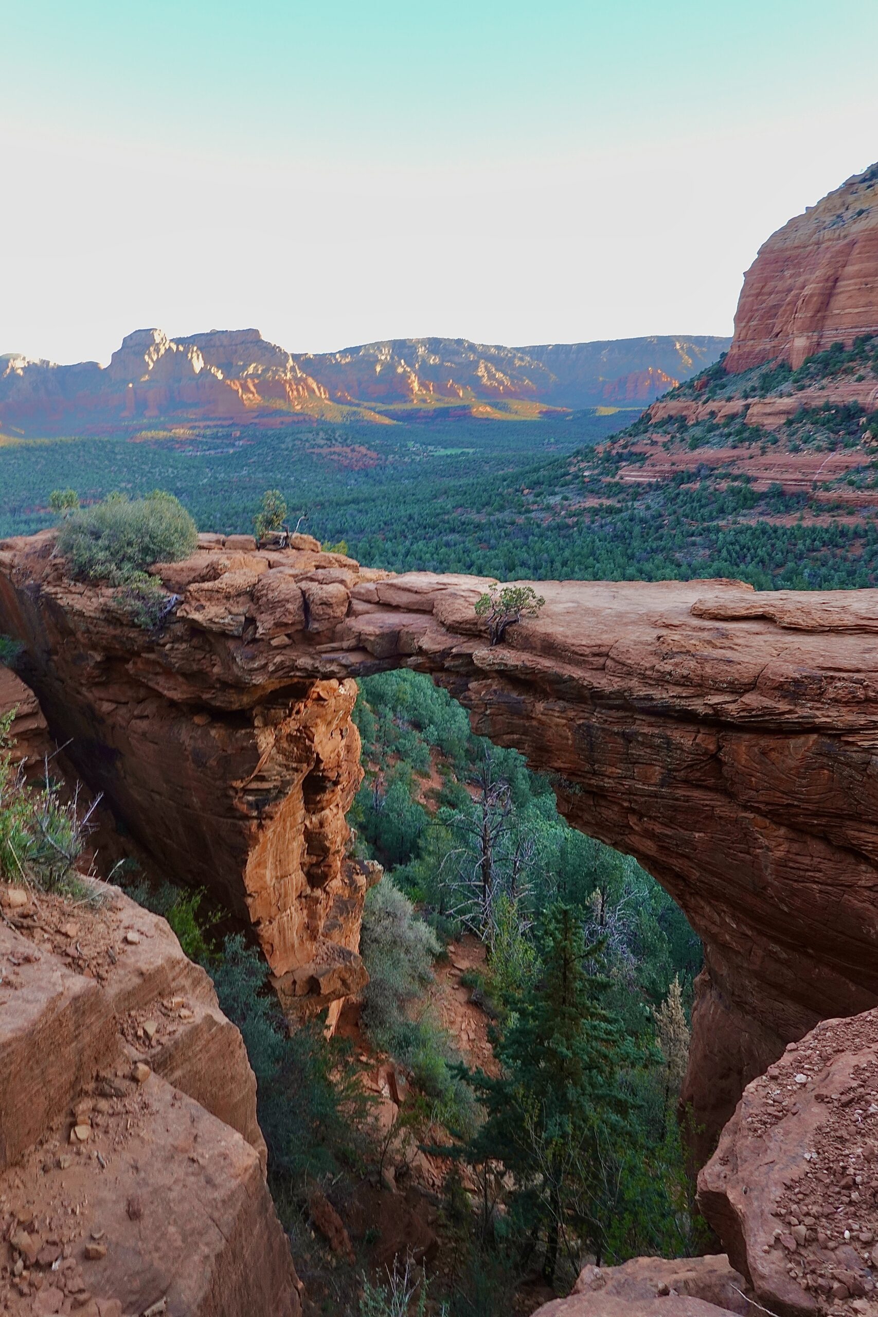 photo of a red rock arch with a green background on a mostly clear day
