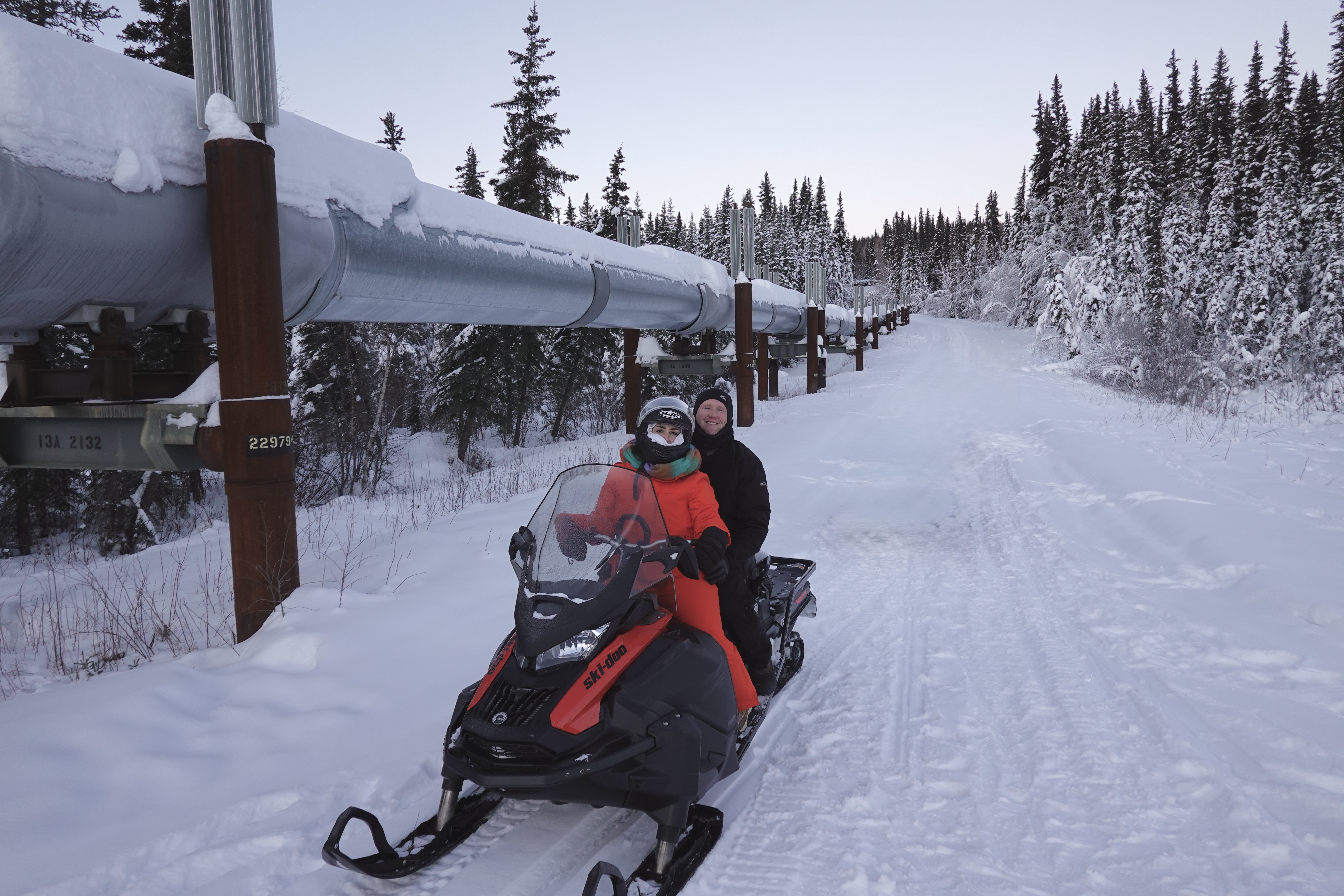 Two people on a snowmobile ride near a snowy pipeline and evergreen trees.