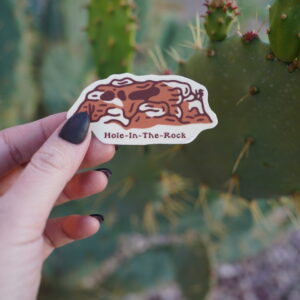 A hand holds a "Hole-In-The-Rock" sticker against a cactus background.