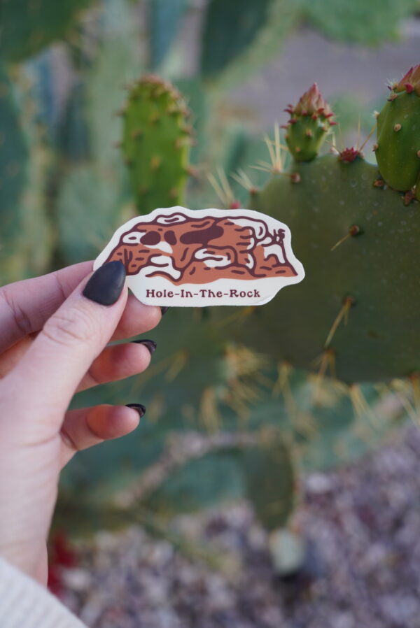 A hand holds a "Hole-In-The-Rock" sticker against a cactus background.