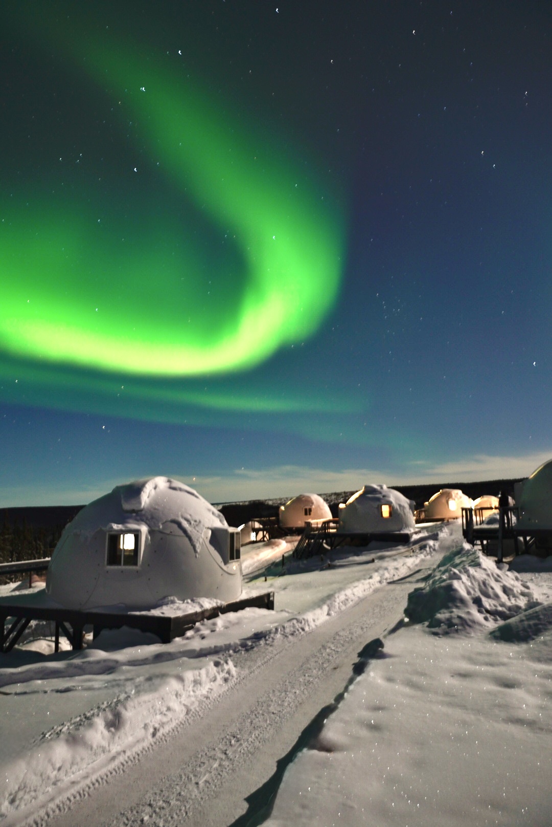 Northern Lights over snow-covered igloo cabins at night.