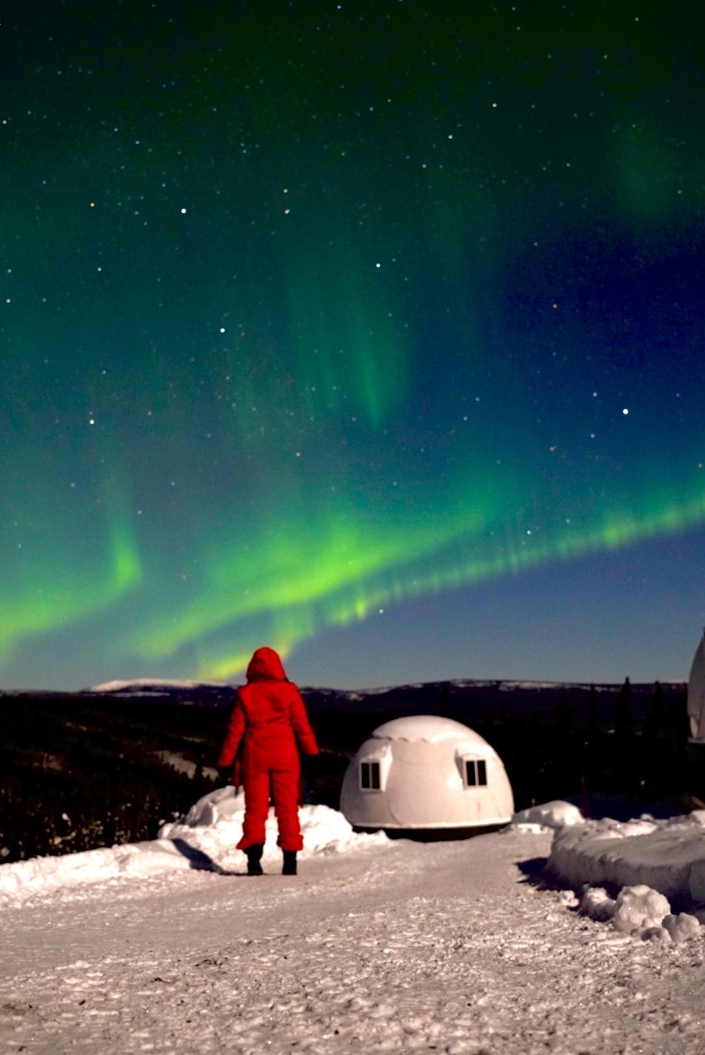 Person in a red snowsuit watching the Northern Lights over snow-covered land with an igloo-shaped building nearby.