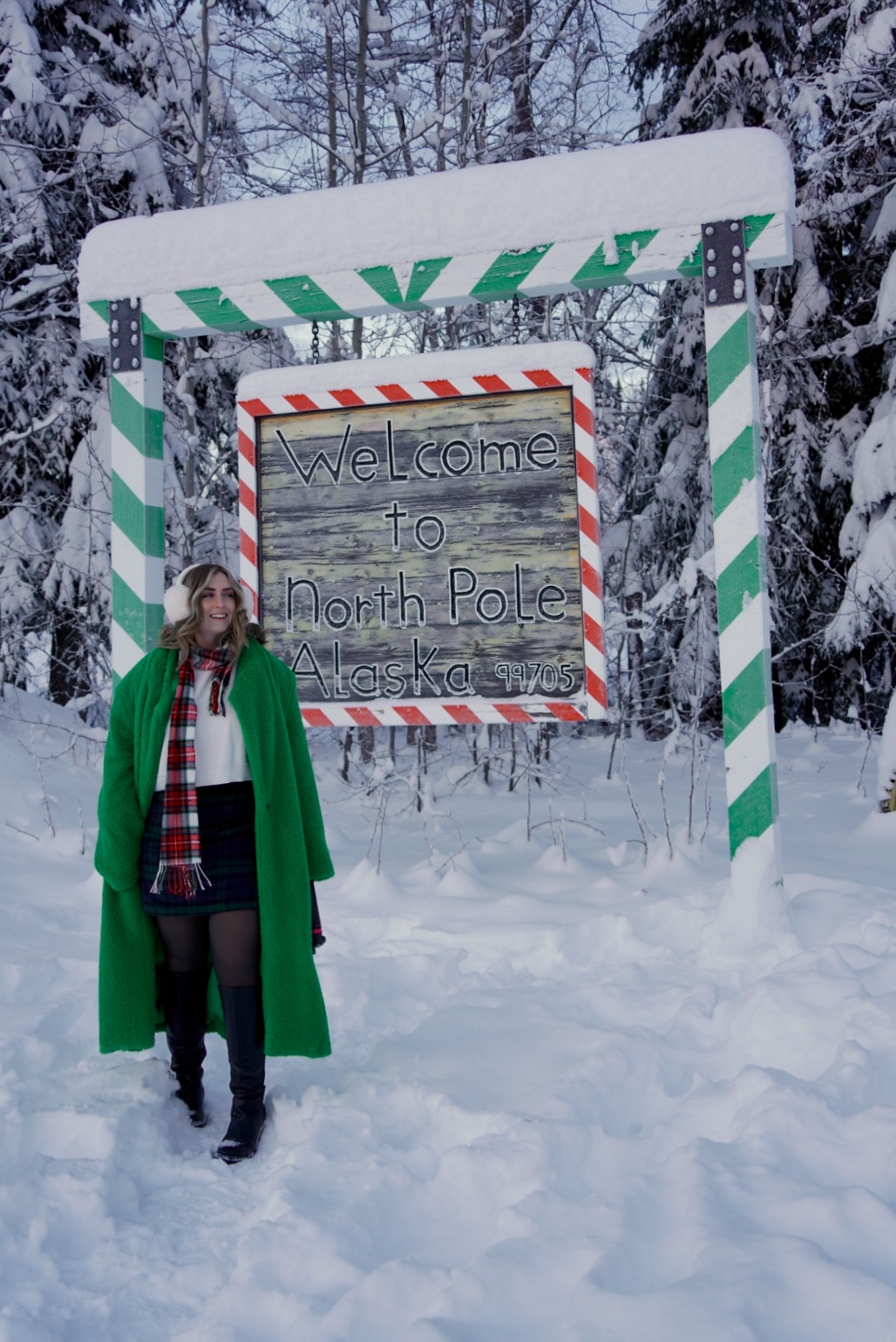 A person in a green coat stands next to a sign reading "Welcome to North Pole, Alaska" amidst a snowy forest.<br />
