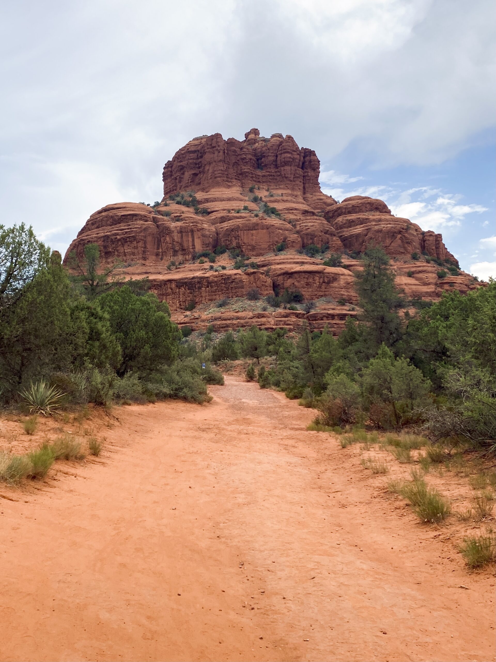 photo of a bell shaped red rock with a clay trail leading up to it and green bushes on each side