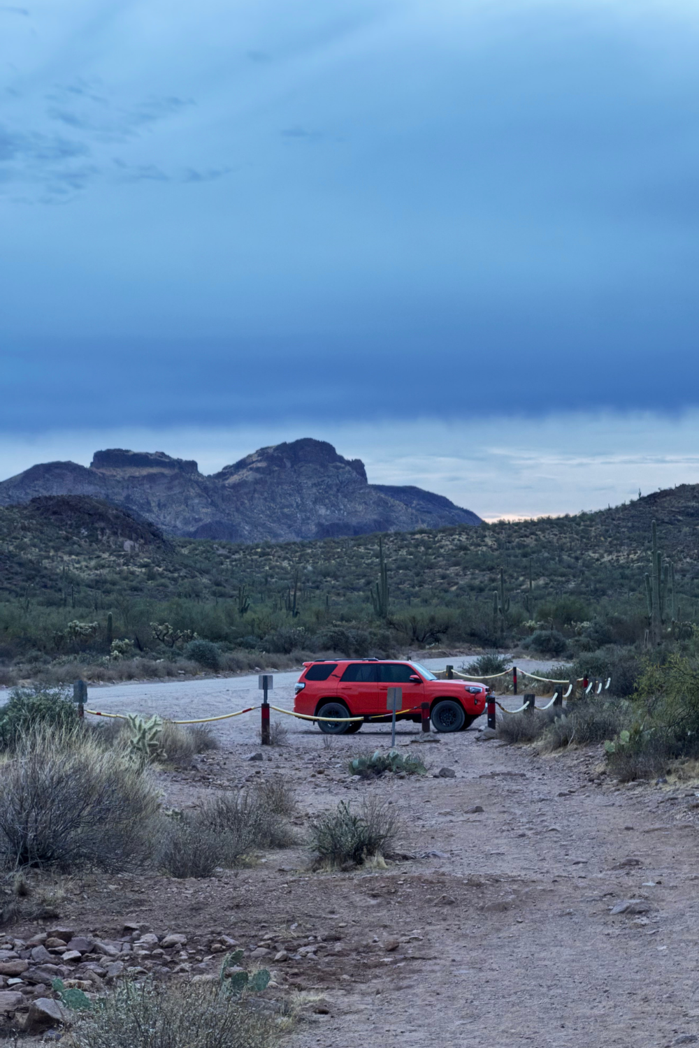 A red SUV parked on a desert road with mountainous terrain and cloudy skies.