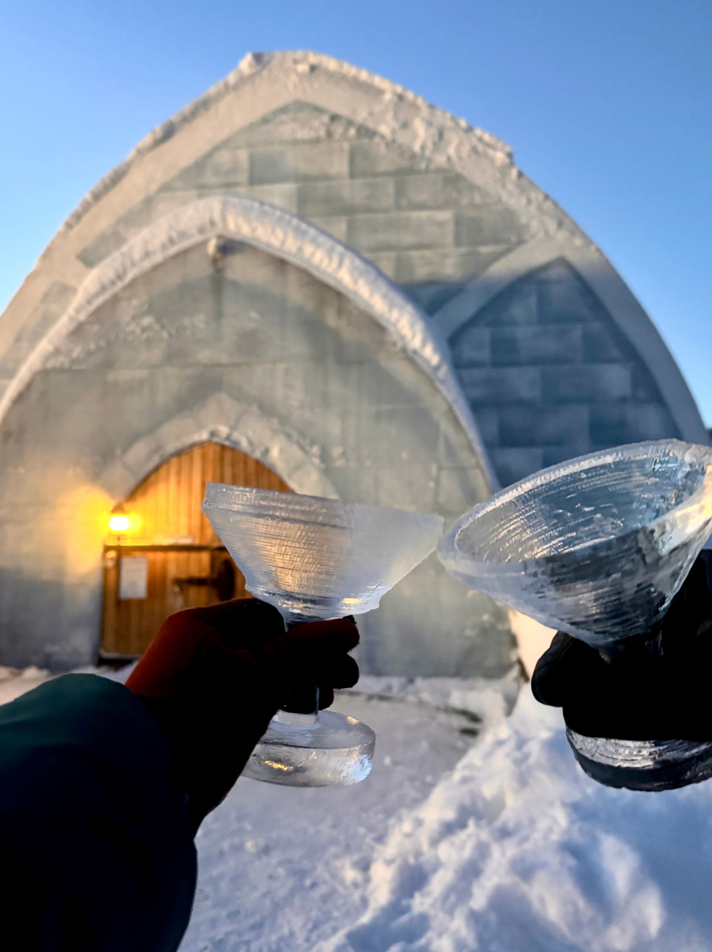 Two gloved hands holding ice goblets in front of an ice building with a wooden door and snow on the ground.