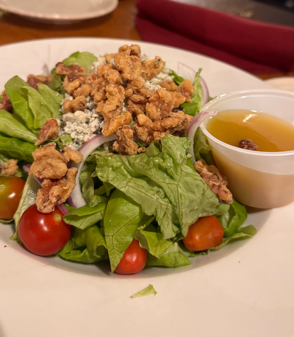 A salad with leafy greens, walnuts, cherry tomatoes, red onions, and crumbled cheese, with a cup of dressing on the side.