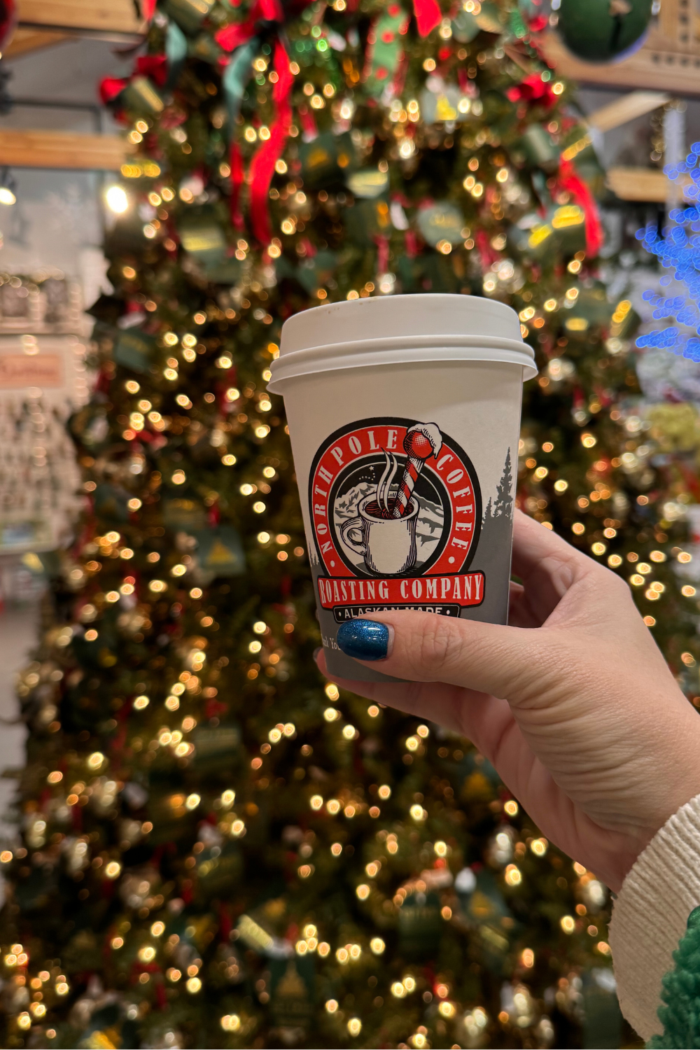 A hand holds a North Pole Coffee cup in front of a decorated Christmas tree.