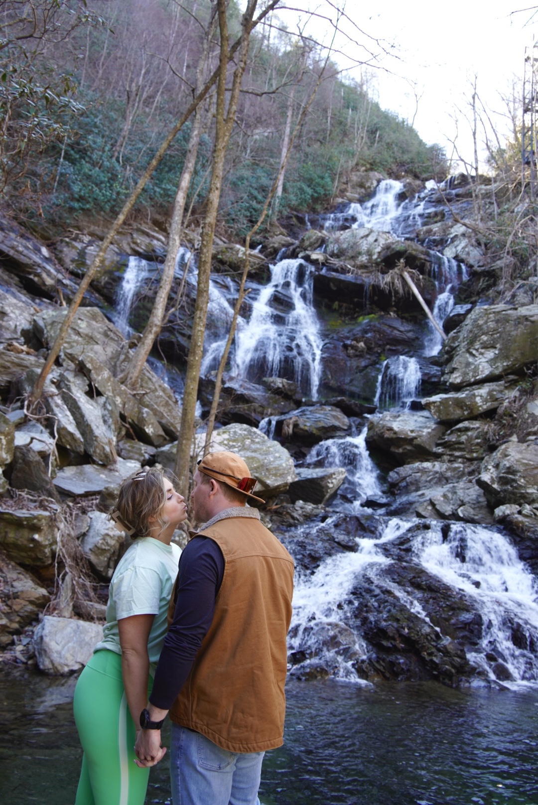 A couple stands in front of a waterfall, surrounded by rocks and trees, about to kiss