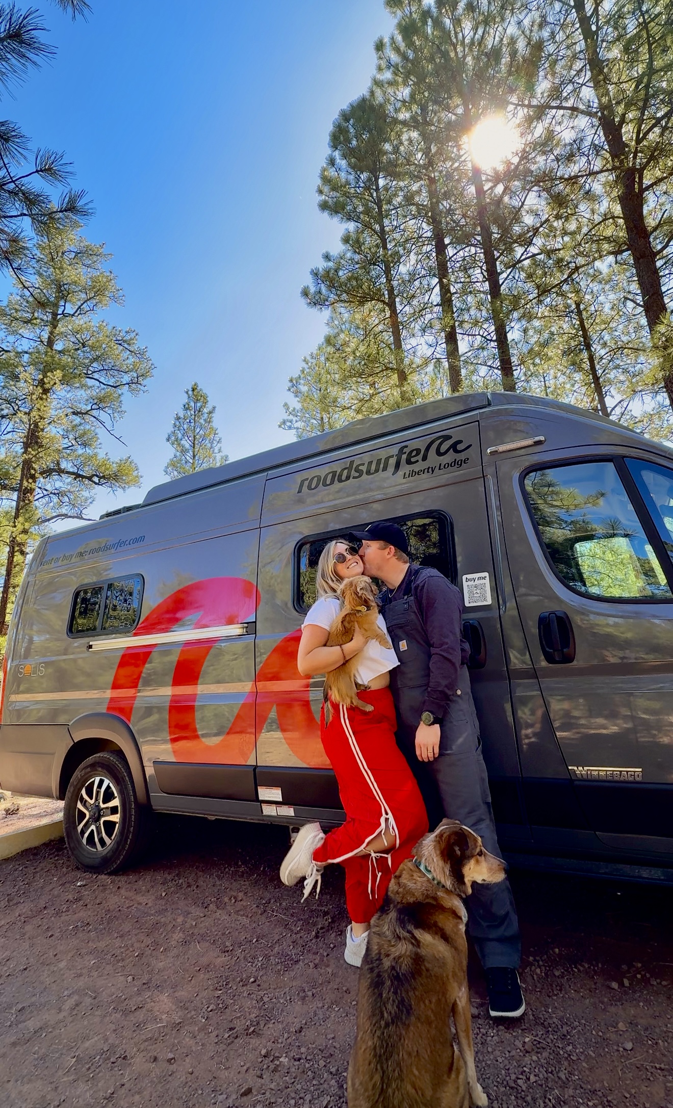 roadsurfer couple with 2 dogs in front of a campervan in the forest in arizona that reads roadsurfer