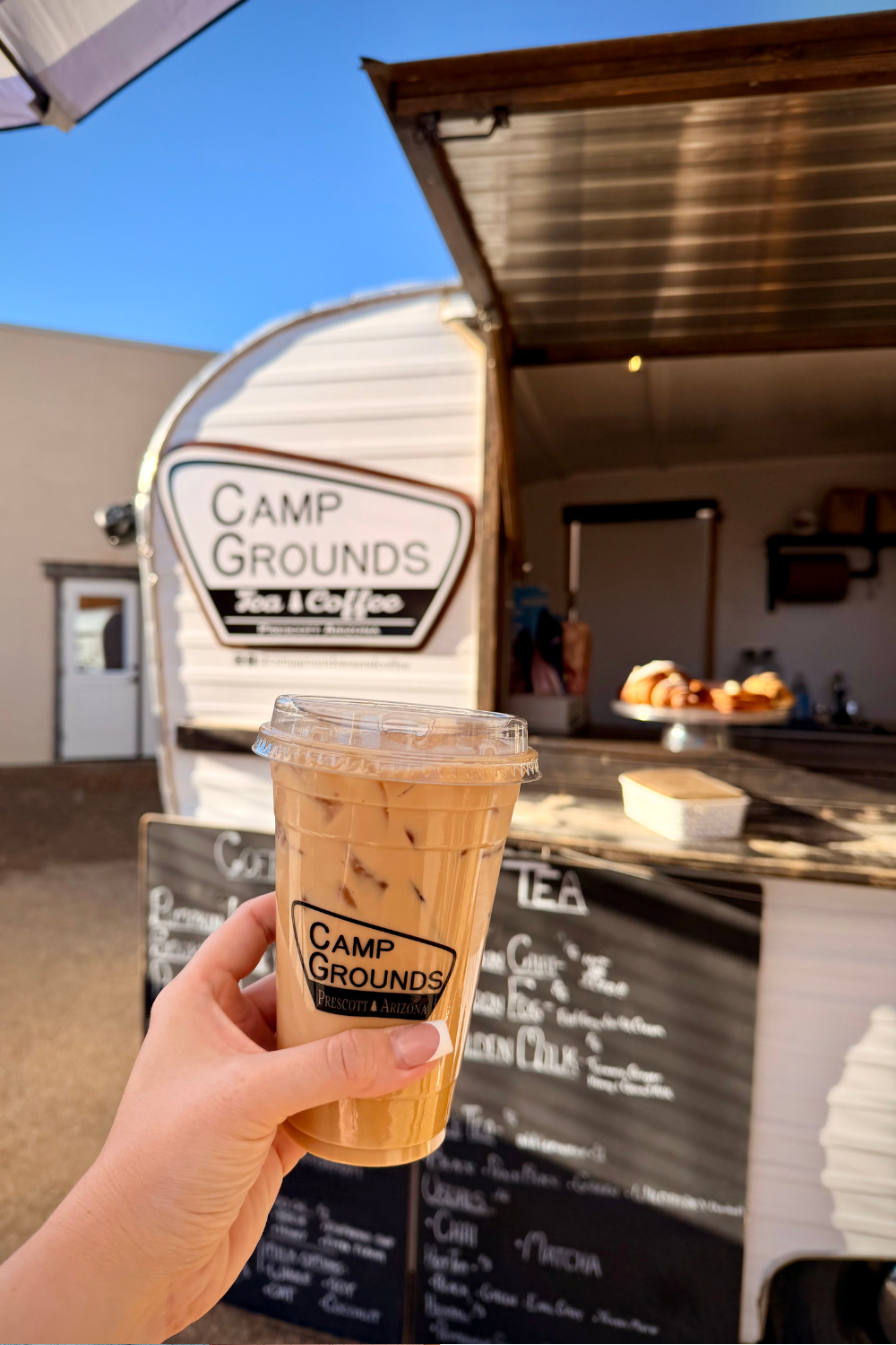 girl holding up an iced latte in front of a sign reading camp grounds
