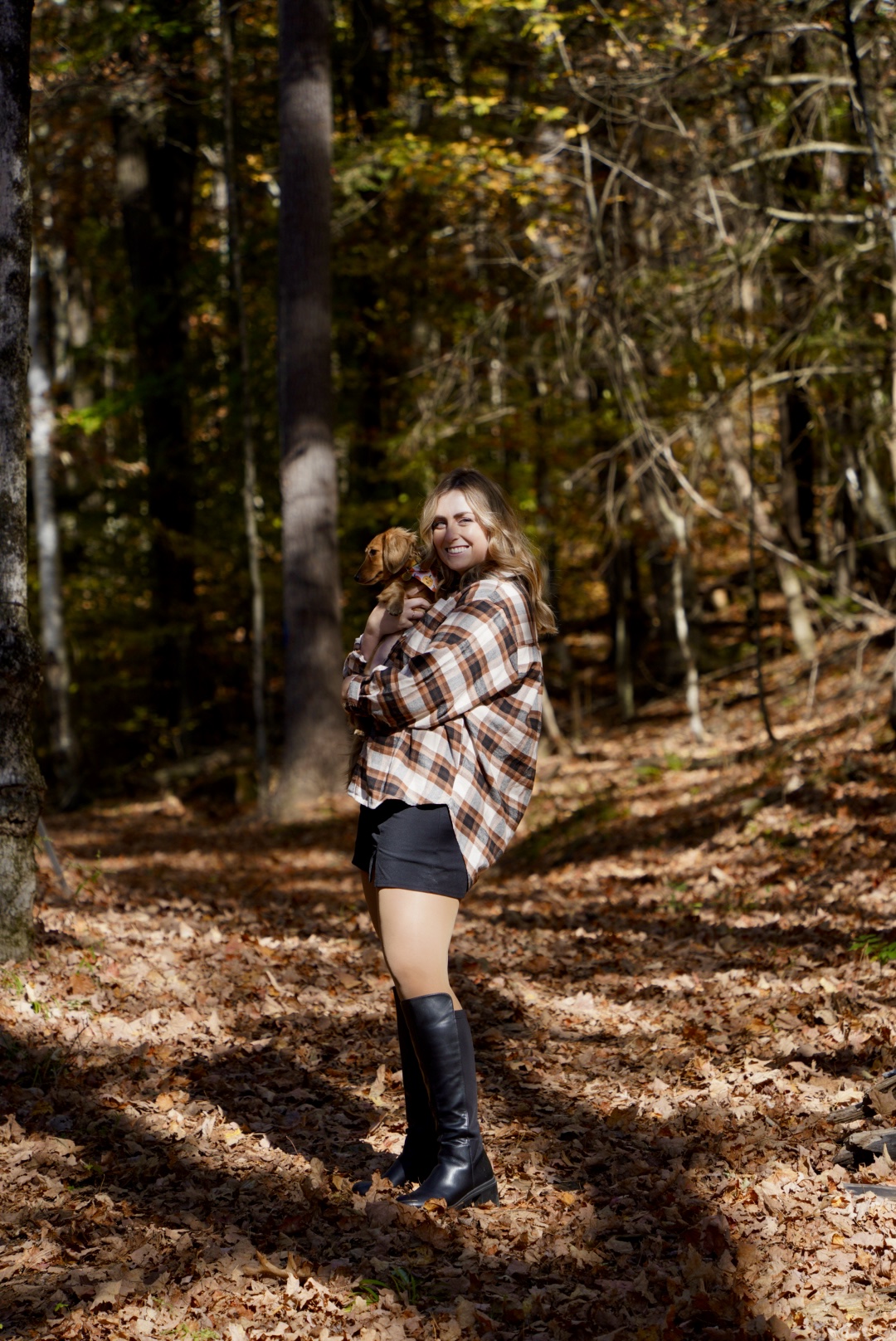 photo of a girl in plaid and a skirt and boots holding a weiner dog in the fall leaves