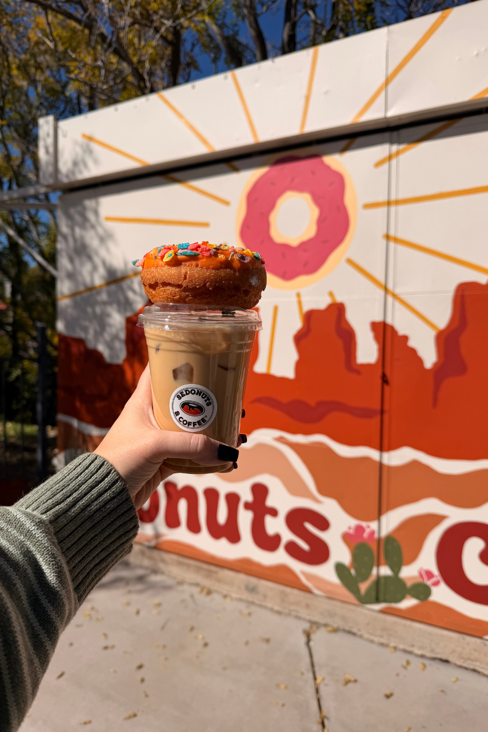 photo of a girl holding up an iced coffee and donut against a mural