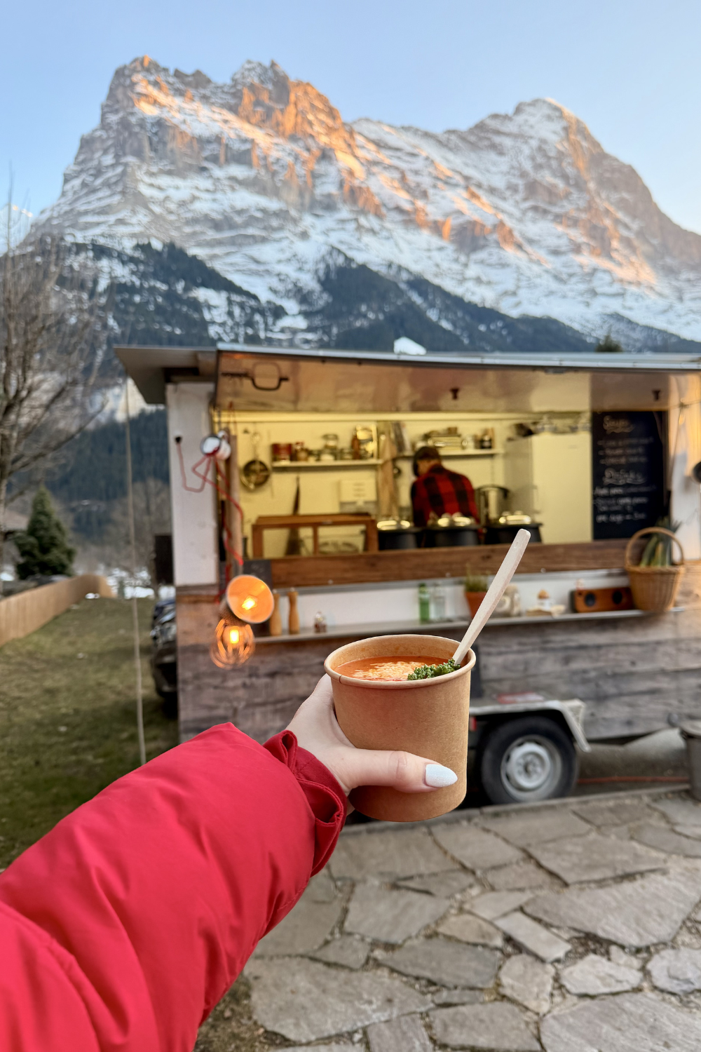photo of a girl holding up a cup of soup in front of a food truck and the mountains