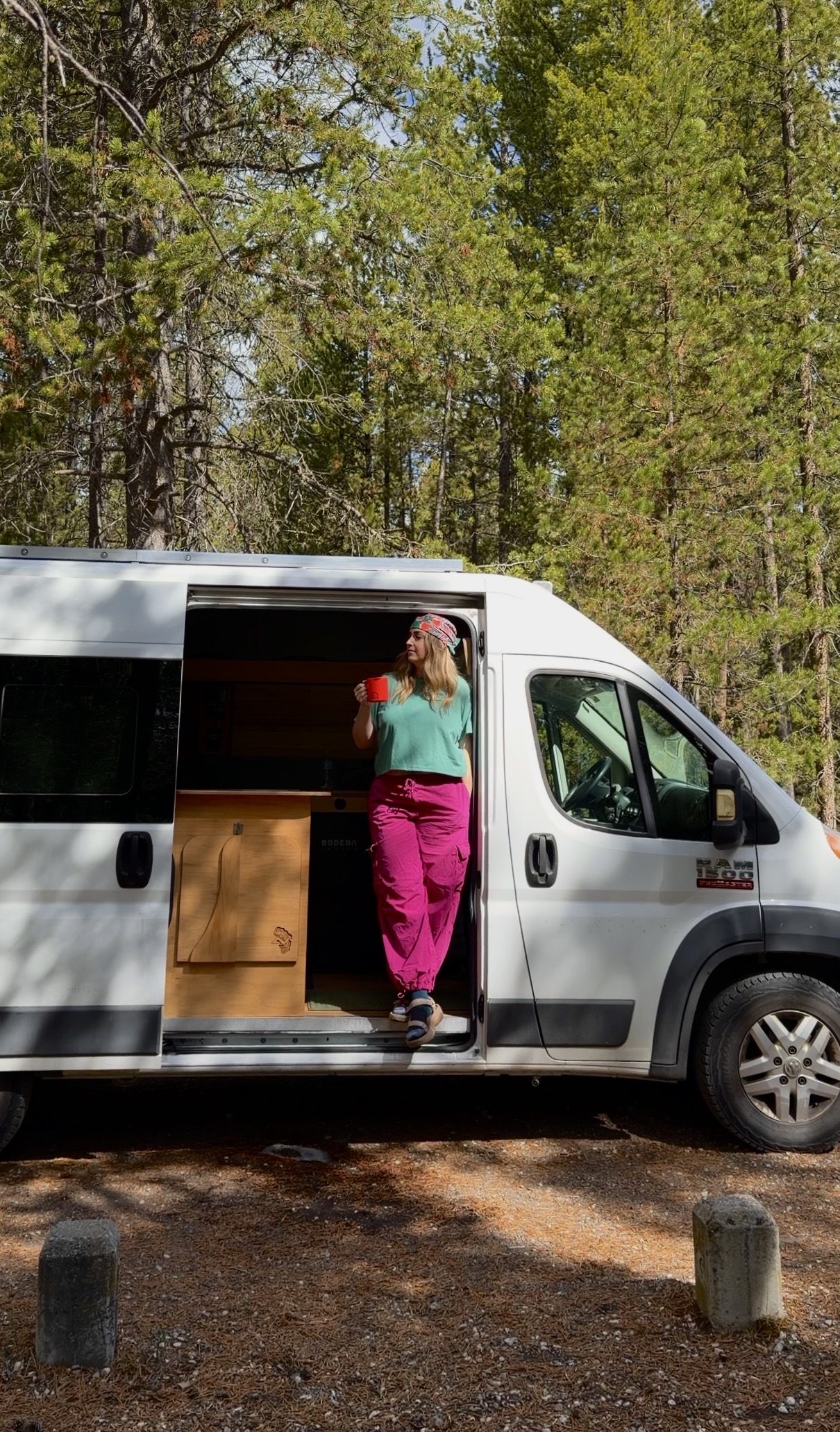 where to stay near yellowstone Woman standing in the open sliding door of a white camper van holding a red coffee mug at a forested campground near Yellowstone National Park