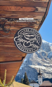 Sign for Eiger Bean Roastery in Grindelwald, Switzerland, with the snow-capped Eiger mountain and village church in the background